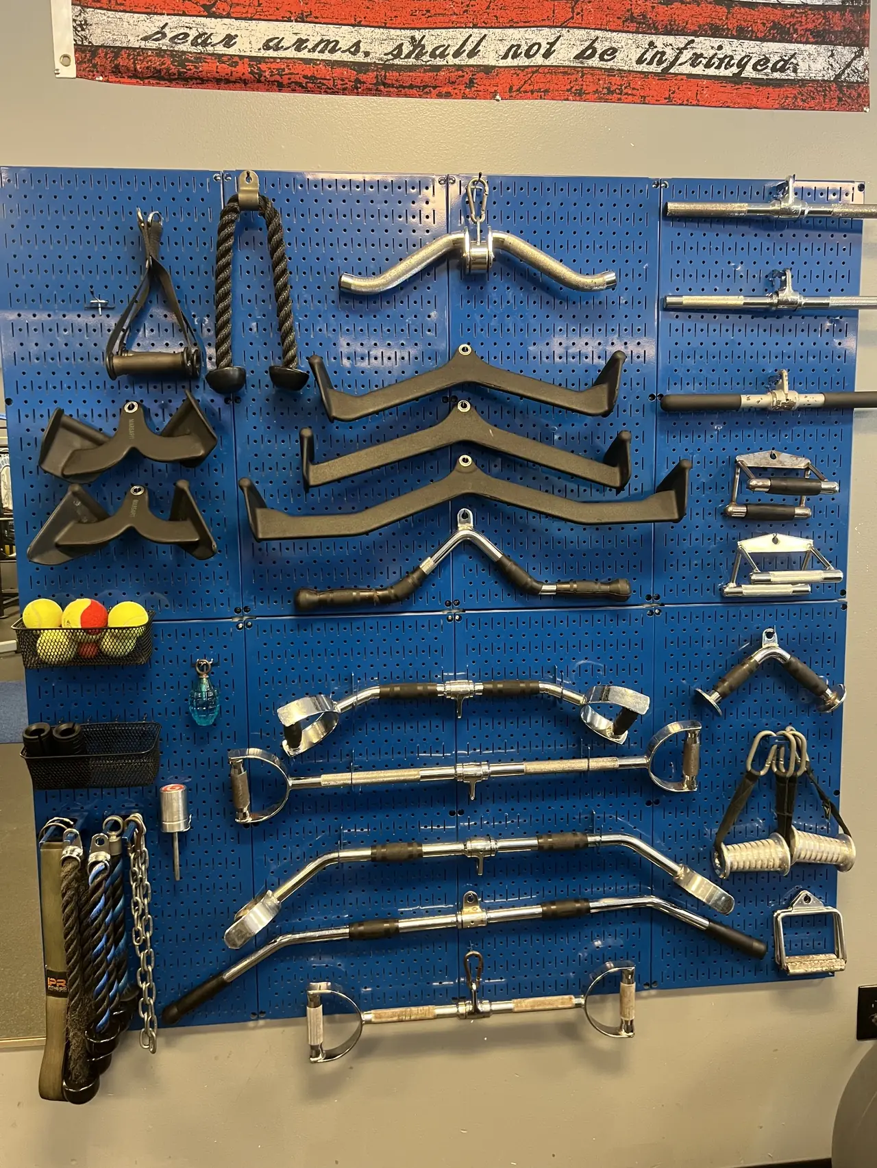 Blue pegboard wall of cable attachments — lat bars, V-handles, tricep ropes, D-handles and straight bars — with battle ropes and a chain stored below.