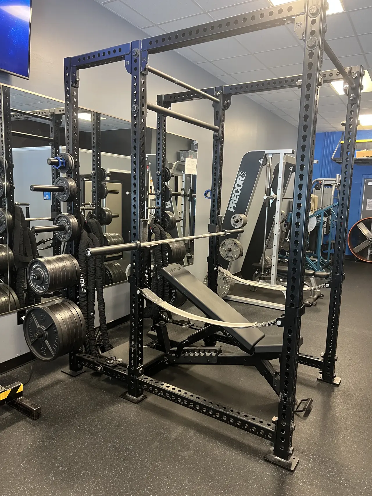 Rogue power rack set up with an adjustable bench and a loaded barbell, plate storage and battle ropes alongside.