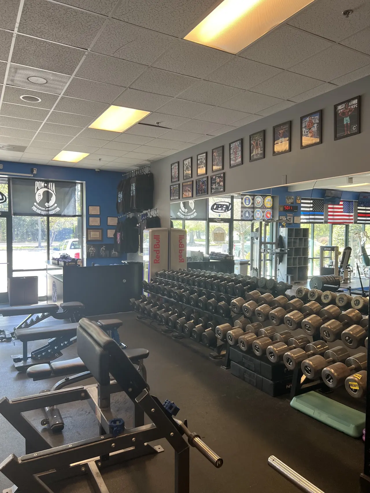 The dumbbell floor seen from the front: full racks, adjustable benches and the wall of framed covers and service flags.